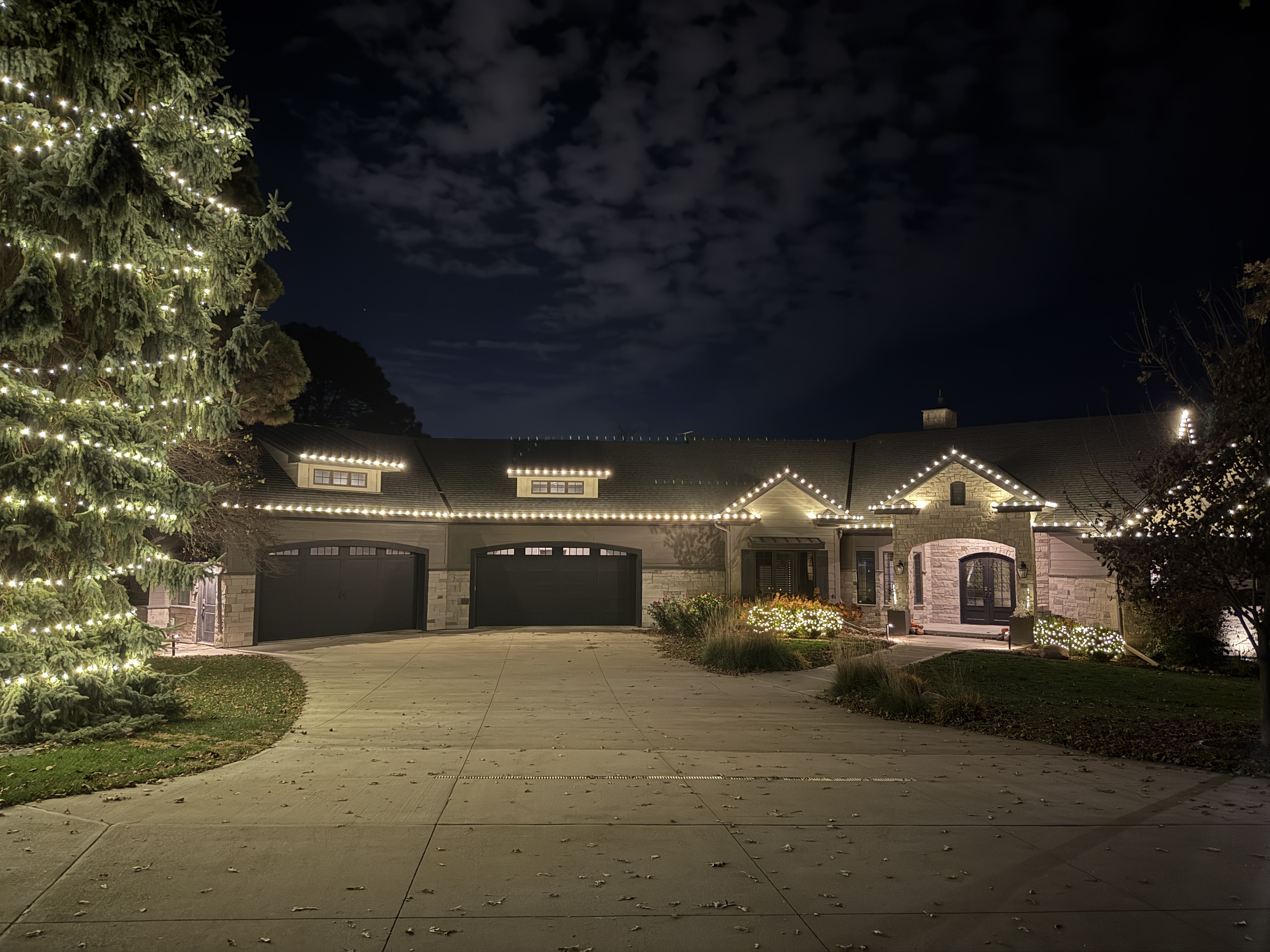 Stone ranch-style home with a warm-white roofline and a towering evergreen wrapped in lights